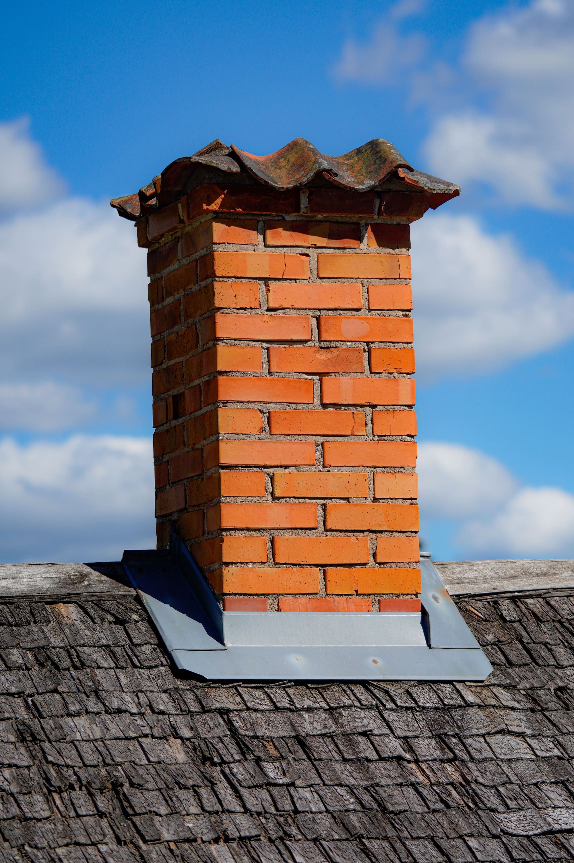 Orange brick chimney with a corbelled top and a pitched tile cap over the flue, with metal apron flashing where it meets a wood shingle roof