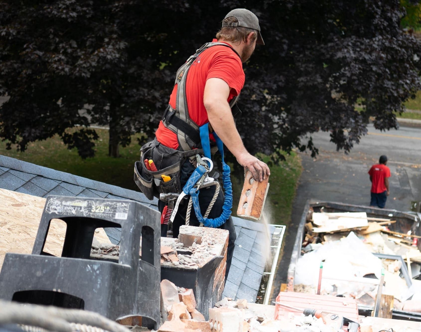 Worker in a fall-arrest harness on a shingle roof taking a brick chimney down by hand, dropping a brick toward a dumpster in the driveway below
