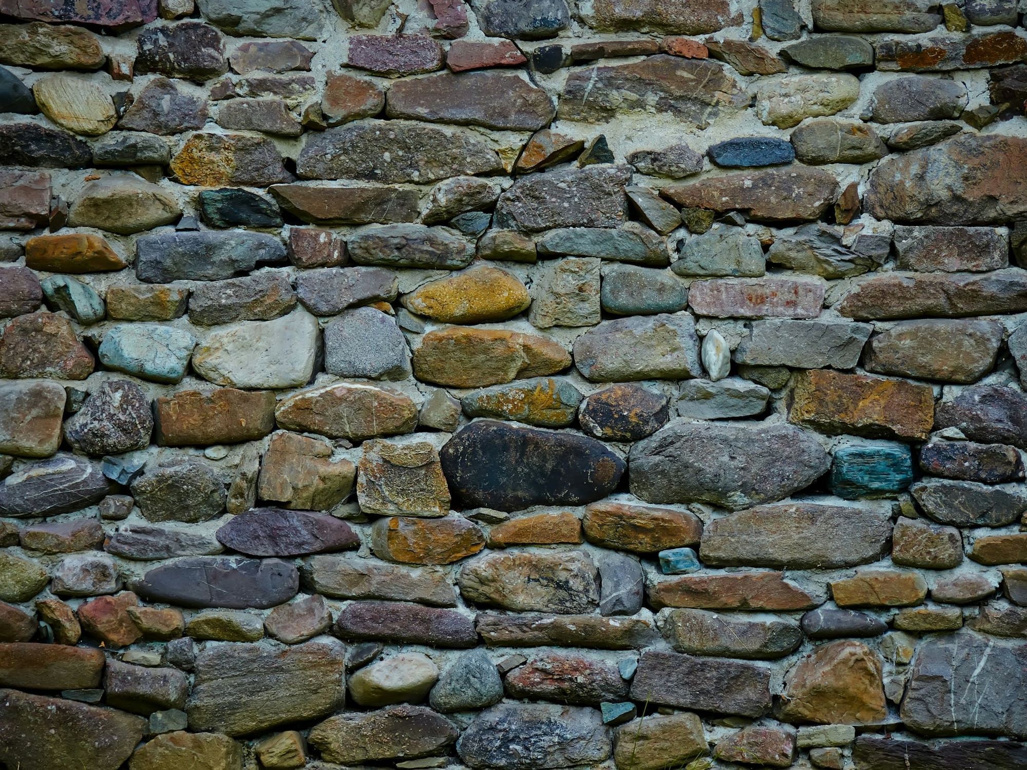 Old wall of rounded fieldstones in mixed grays, greens and browns, laid in rough courses with weathered mortar